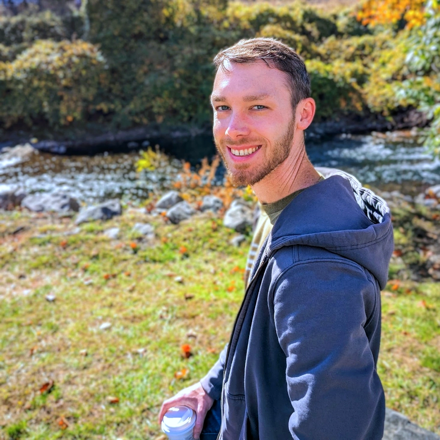 a photo of Nick dressed in a blue hoodie holding a coffee. He's standing by a river on a sunny autumn day.
